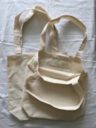 Two beige cloth bags with handles laid flat on a slightly crumpled fabric surface. The bags have a simple, unadorned design and appear to be made of cotton or canvas material.