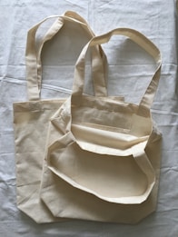 Two beige cloth bags with handles laid flat on a slightly crumpled fabric surface. The bags have a simple, unadorned design and appear to be made of cotton or canvas material.