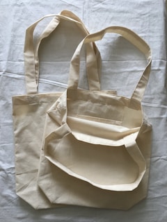 Two beige cloth bags with handles laid flat on a slightly crumpled fabric surface. The bags have a simple, unadorned design and appear to be made of cotton or canvas material.