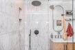 A modern bathroom with marble walls and a sleek, black showerhead. The shower area features built-in niches for toiletries. Outside the shower, there is a circular mirror, a wooden vanity with a white countertop, and a red towel hanging nearby. A wooden bath brush is seen hanging inside the shower enclosure.