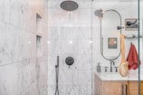 A modern bathroom with marble walls and a sleek, black showerhead. The shower area features built-in niches for toiletries. Outside the shower, there is a circular mirror, a wooden vanity with a white countertop, and a red towel hanging nearby. A wooden bath brush is seen hanging inside the shower enclosure.