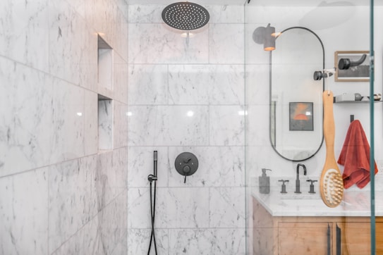 A modern bathroom with marble walls and a sleek, black showerhead. The shower area features built-in niches for toiletries. Outside the shower, there is a circular mirror, a wooden vanity with a white countertop, and a red towel hanging nearby. A wooden bath brush is seen hanging inside the shower enclosure.