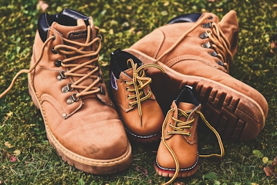 Brown leather adult work boots are placed on green grass. In front of them, two smaller, similar style brown leather boots intended for children are positioned. The boots have sturdy soles and are laced with yellow laces.
