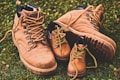 Brown leather adult work boots are placed on green grass. In front of them, two smaller, similar style brown leather boots intended for children are positioned. The boots have sturdy soles and are laced with yellow laces.