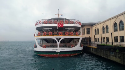 A ferry docked by a building with arched windows along a waterfront. The ferry has multiple decks with orange life rings on the rails and a Turkish flag at the top. The water appears choppy under an overcast sky, and there are a few birds visible in the air and water.
