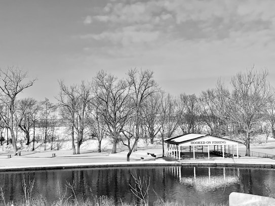 A serene winter scene with a pond in the foreground and a wooden pavilion labeled 'Hooked on Fishing' by the water. Bare trees are spread throughout the snowy landscape, and a clear sky with a few clouds is overhead.