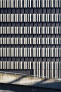 A facade of a building featuring a repetitive pattern of vertical metallic panels that create a wavy texture across the surface. The panels reflect light and cast shadows, adding depth and visual interest to the structure. Concrete elements are visible at the bottom, contributing to an industrial aesthetic.