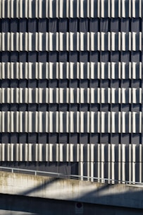 A facade of a building featuring a repetitive pattern of vertical metallic panels that create a wavy texture across the surface. The panels reflect light and cast shadows, adding depth and visual interest to the structure. Concrete elements are visible at the bottom, contributing to an industrial aesthetic.