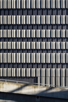 A facade of a building featuring a repetitive pattern of vertical metallic panels that create a wavy texture across the surface. The panels reflect light and cast shadows, adding depth and visual interest to the structure. Concrete elements are visible at the bottom, contributing to an industrial aesthetic.
