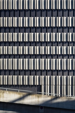 A facade of a building featuring a repetitive pattern of vertical metallic panels that create a wavy texture across the surface. The panels reflect light and cast shadows, adding depth and visual interest to the structure. Concrete elements are visible at the bottom, contributing to an industrial aesthetic.