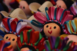 Several doll faces are adorned with colorful ribbons. The dolls have smiling expressions with simple, round, black eyes and red geometric mouths. The ribbons are arranged in vibrant patterns, creating a festive and lively appearance.
