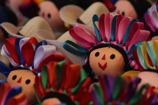Several doll faces are adorned with colorful ribbons. The dolls have smiling expressions with simple, round, black eyes and red geometric mouths. The ribbons are arranged in vibrant patterns, creating a festive and lively appearance.