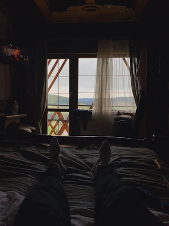 A person is lounging on a bed with feet outstretched, facing a window that overlooks a scenic landscape. Curtains are partially drawn, allowing a view of distant mountains and trees. The room is dimly lit, creating a cozy and tranquil atmosphere.