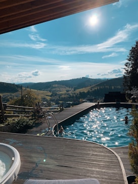 A serene outdoor pool with a deck sits overlooking a scenic valley, basked in sunlight. The crystal-clear water reflects the brightness of the day as a few people enjoy a swim. Rolling hills and a bright blue sky with scattered clouds provide a picturesque background. A section of a wooden structure, possibly a roof, is visible at the top.