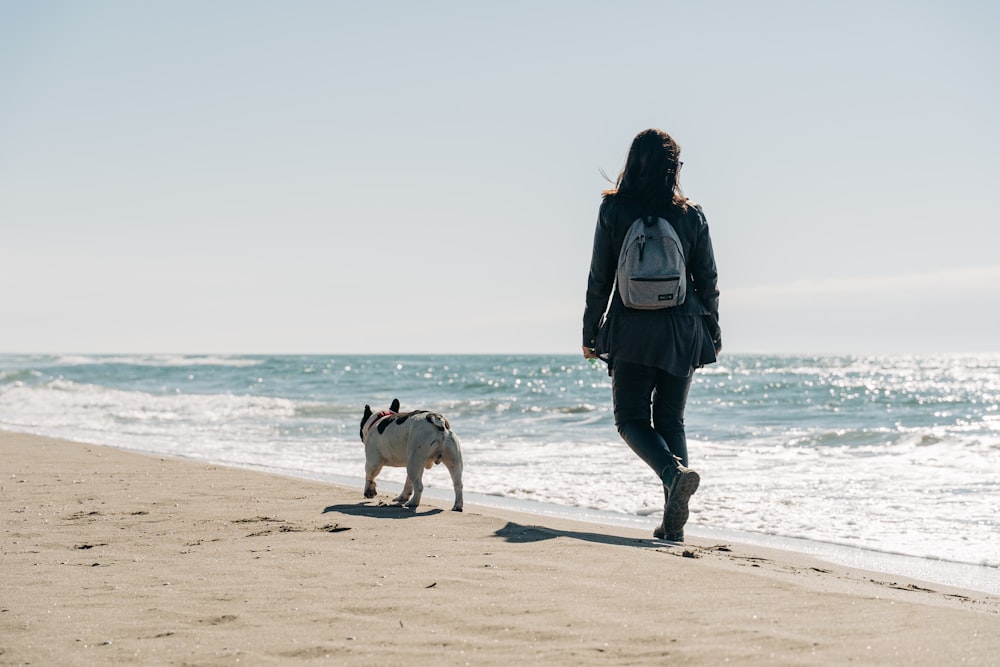 Mujer en chaqueta negra caminando en la playa con perro blanco durante el día
