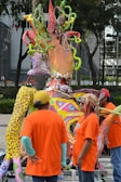 A vibrant and colorful sculpture with organic shapes stands outdoors surrounded by people wearing bright orange shirts. The sculpture contains twisted, multicolored elements resembling tentacles or vines. The people appear to be observing or engaging with the artwork in a casual, outdoor setting with trees and a building in the background.