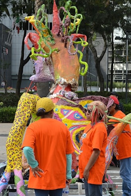 A vibrant and colorful sculpture with organic shapes stands outdoors surrounded by people wearing bright orange shirts. The sculpture contains twisted, multicolored elements resembling tentacles or vines. The people appear to be observing or engaging with the artwork in a casual, outdoor setting with trees and a building in the background.