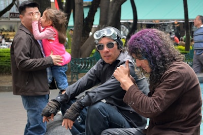 A man is holding and playing with a child, while two men are sitting on a bench. One of the men sitting has colored hair and is adjusting sunglasses, while the other is wearing goggles on his forehead. Trees and a green canopy are visible in the background along with other people.