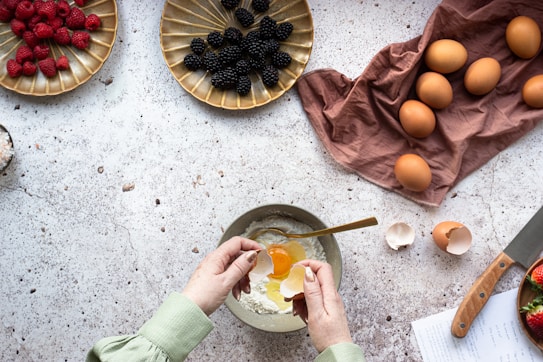 A person is cracking an egg into a bowl surrounded by various ingredients on a textured countertop. To the left, raspberries are arranged on a golden plate, while blackberries are on another similar plate above. Several eggs rest on a brown cloth, and a wooden-handled knife lies nearby. Eggshells are scattered around, and a piece of paper with handwritten notes can be seen beside a few strawberries.