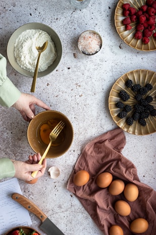 A person is preparing a baking mixture with a bowl of eggs and a fork. Other ingredients include flour, a bowl of salt, raspberries, and blackberries arranged on distinct plates. Several whole eggs are placed on a brown cloth. A knife rests nearby, and a page with handwritten notes can be seen partially.