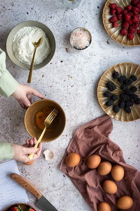 A person is preparing a baking mixture with a bowl of eggs and a fork. Other ingredients include flour, a bowl of salt, raspberries, and blackberries arranged on distinct plates. Several whole eggs are placed on a brown cloth. A knife rests nearby, and a page with handwritten notes can be seen partially.