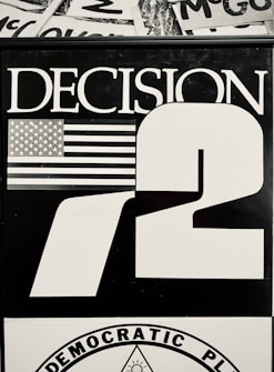 A black and white graphic featuring the word 'DECISION' in large capital letters next to the number '72'. An illustration of the American flag is present beside the word 'DECISION'. The bottom part includes a circular emblem with the words 'DEMOCRATIC PLATFORM' around it.