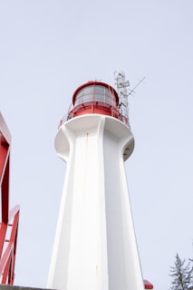 A tall white lighthouse with a red top section, featuring a metal antenna or broadcasting structure attached to the side. The structure is photographed from a low angle, emphasizing its height against a pale sky.