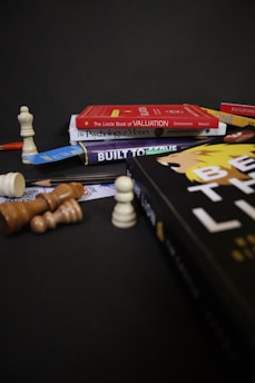 A collection of books is stacked against a dark background, including titles like 'The Little Book of Valuation,' 'The Psychology of Money,' and 'Built to Thrive.' A chessboard setup with various chess pieces is arranged around the books, some standing and others toppled over.