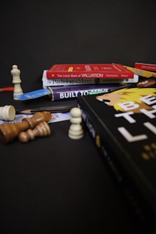 A collection of books is stacked against a dark background, including titles like 'The Little Book of Valuation,' 'The Psychology of Money,' and 'Built to Thrive.' A chessboard setup with various chess pieces is arranged around the books, some standing and others toppled over.