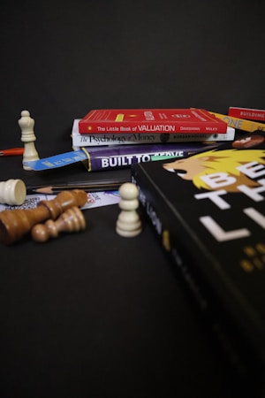 A collection of books is stacked against a dark background, including titles like 'The Little Book of Valuation,' 'The Psychology of Money,' and 'Built to Thrive.' A chessboard setup with various chess pieces is arranged around the books, some standing and others toppled over.