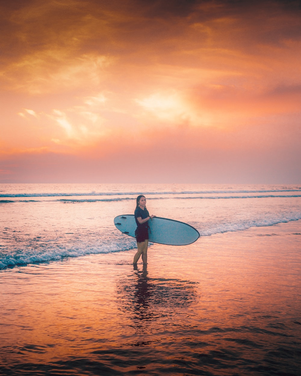 homem na camisa branca segurando a prancha de surf branca andando na praia durante o pôr do sol