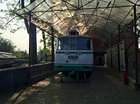 A vintage, pale green and white bus is parked under an open, metal-framed canopy surrounded by trees. The setting appears to be historical or ceremonial, with informational panels lining the side of the area. The scene suggests a peaceful outdoor environment with natural elements.