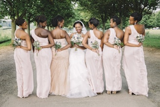 A group of women in formal dresses stand together outdoors. They are dressed similarly with bouquets in hand. The central figure appears to be a bride, wearing a white gown and veil. The setting is a tree-lined path, suggesting a park or outdoor venue.