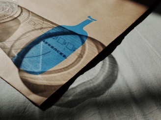 A cardboard or paper surface with a printed blue bottle shape casts a shadow alongside a transparent plastic cup. The shadow adds depth and dimension, and the lighting creates a play of light and dark on the surface.