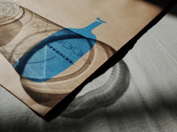 A cardboard or paper surface with a printed blue bottle shape casts a shadow alongside a transparent plastic cup. The shadow adds depth and dimension, and the lighting creates a play of light and dark on the surface.