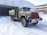 A vintage military-style truck with a rugged design is parked on a snow-covered surface. The vehicle features large tires, a green wooden cargo area, and a red front covering. It is situated near an industrial building with a weathered appearance, which has an open garage area.