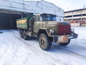 A vintage military-style truck with a rugged design is parked on a snow-covered surface. The vehicle features large tires, a green wooden cargo area, and a red front covering. It is situated near an industrial building with a weathered appearance, which has an open garage area.