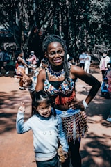 A woman dressed in traditional, colorful attire with beaded jewelry and face paint stands next to a smiling child in casual clothing. They are both gesturing with their hands, possibly indicating peace signs, in an outdoor setting with other people in the background.