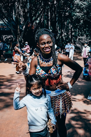 A woman dressed in traditional, colorful attire with beaded jewelry and face paint stands next to a smiling child in casual clothing. They are both gesturing with their hands, possibly indicating peace signs, in an outdoor setting with other people in the background.