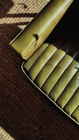 A green upholstered chair with a smooth, textured surface is partially visible on the edge of a brown carpeted floor. The lighting creates shadows and highlights on both the seat and the carpet, adding depth to the textures.