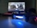 A video game controller with a glowing blue light sits on a table in front of a computer screen displaying a racing game, featuring a detailed view of a race car.