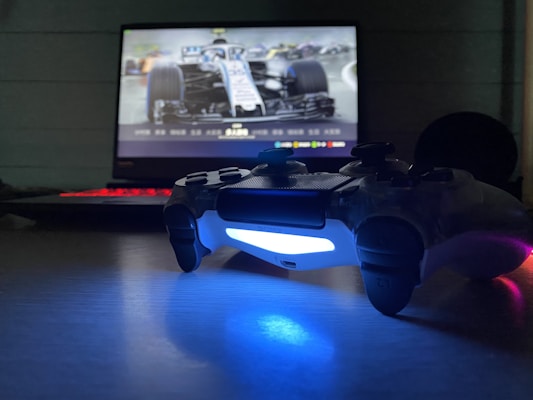 A video game controller with a glowing blue light sits on a table in front of a computer screen displaying a racing game, featuring a detailed view of a race car.