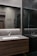 A modern bathroom interior with sleek, dark marble walls and a large mirror that spans the countertop area. The sink is built into a wooden vanity, and there is a chrome faucet. Subtle lighting is embedded above the mirror, casting a soft glow around the room.