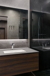 A modern bathroom interior with sleek, dark marble walls and a large mirror that spans the countertop area. The sink is built into a wooden vanity, and there is a chrome faucet. Subtle lighting is embedded above the mirror, casting a soft glow around the room.