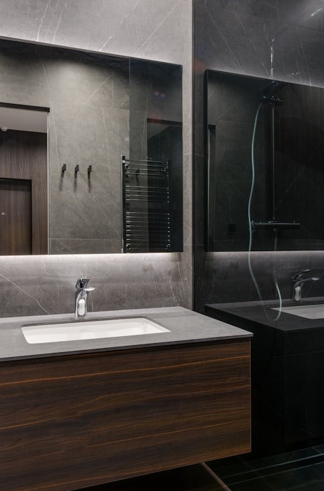 A modern bathroom interior with sleek, dark marble walls and a large mirror that spans the countertop area. The sink is built into a wooden vanity, and there is a chrome faucet. Subtle lighting is embedded above the mirror, casting a soft glow around the room.