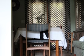A dining area with a wooden table and four chairs, two featuring plaid upholstery and one with a cloth draped over it. A small plant is placed on the table, which is covered with a white tablecloth. The backdrop includes patterned curtains that allow some light to filter through, creating a cozy atmosphere. To the right, there is a cabinet or sideboard.
