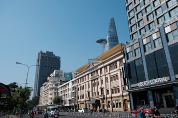 A bustling urban street scene with modern and traditional architecture. The foreground features a busy road with cars and motorbikes. To the right, a tall, modern building with a distinctive conical top stands prominently against a clear blue sky, alongside older, ornate buildings. Signs indicating the names of businesses, including one that says 'Liberty Central,' are visible on the buildings.
