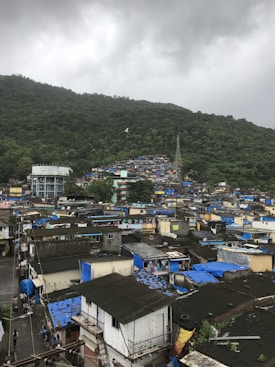 A densely packed settlement with numerous small houses, many of which have blue tarpaulin roofs. The area is surrounded by lush green hills under a cloudy sky, creating a contrast between the green landscape and the urban sprawl.
