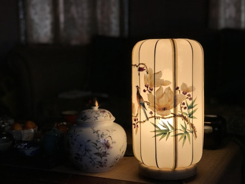 A beautifully decorated cylindrical lamp emits a warm, soft glow. The lamp features intricate floral designs with bird motifs, adding a touch of elegance to the surrounding darkness. Beside the lamp, a porcelain jar with floral patterns complements the aesthetic, creating a harmonious and peaceful setting.