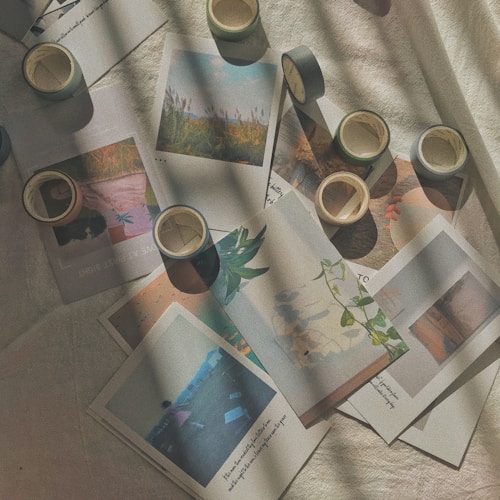 A collection of printed photographs scattered on a textured fabric surface, with rolls of washi tape interspersed among them. The photos show various scenes, mostly nature-related, and are softly lit with shadow patterns cast across them, adding a serene and nostalgic feel.
